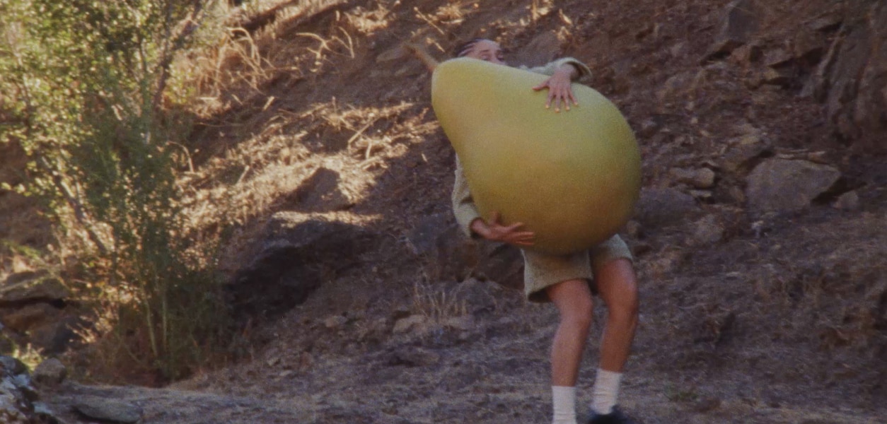 Video of Adwoa Aboah carrying around a large pear fruit prop along a dry, rocky, hillside path.